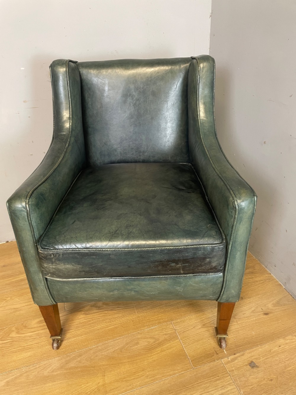 An Edwardian green leather armchair with sloping sides, a square seat cushion, and square tapered mahogany legs fitted with brass castors. View VII