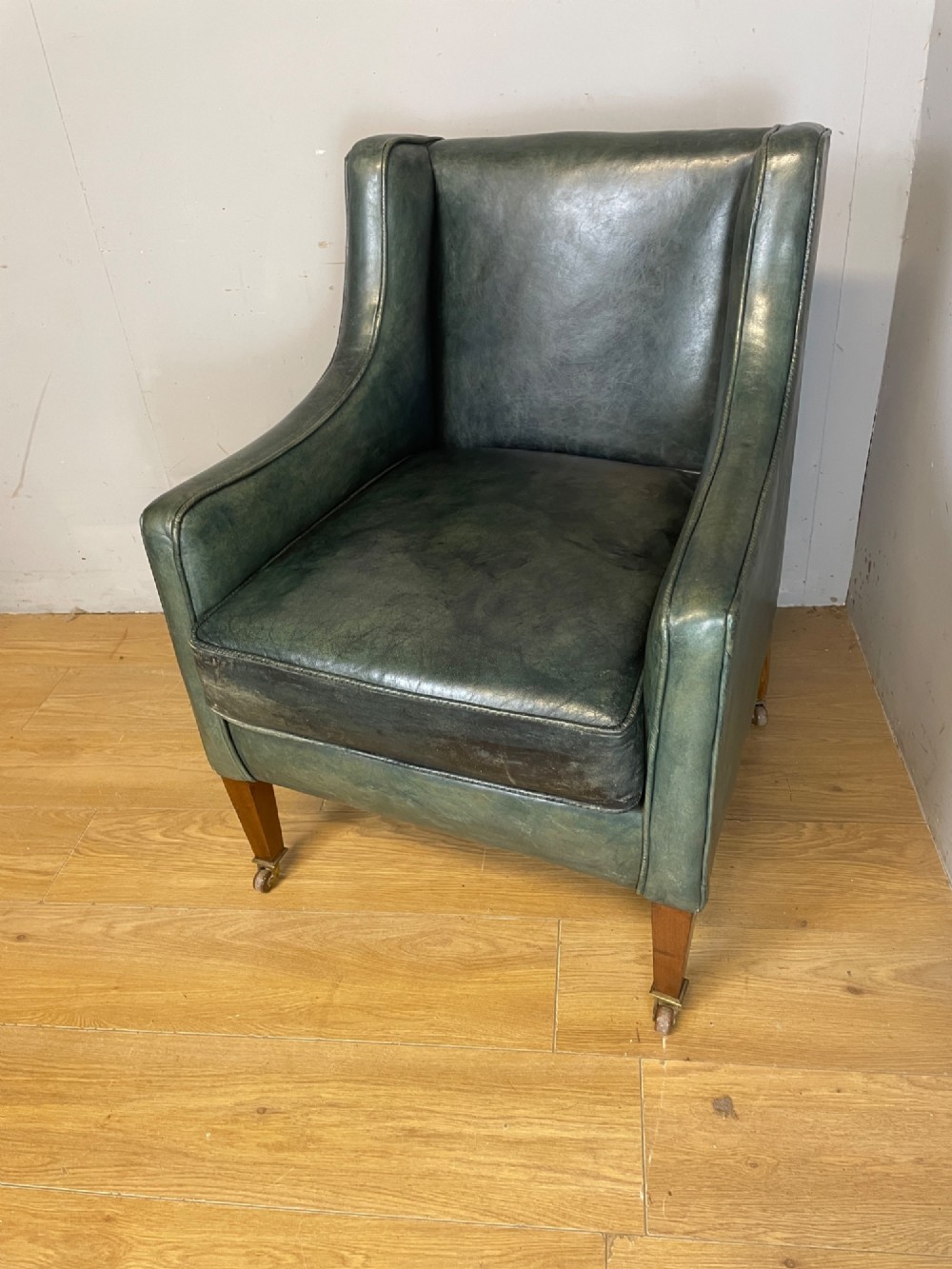 An Edwardian green leather armchair with sloping sides, a square seat cushion, and square tapered mahogany legs fitted with brass castors. View VIII