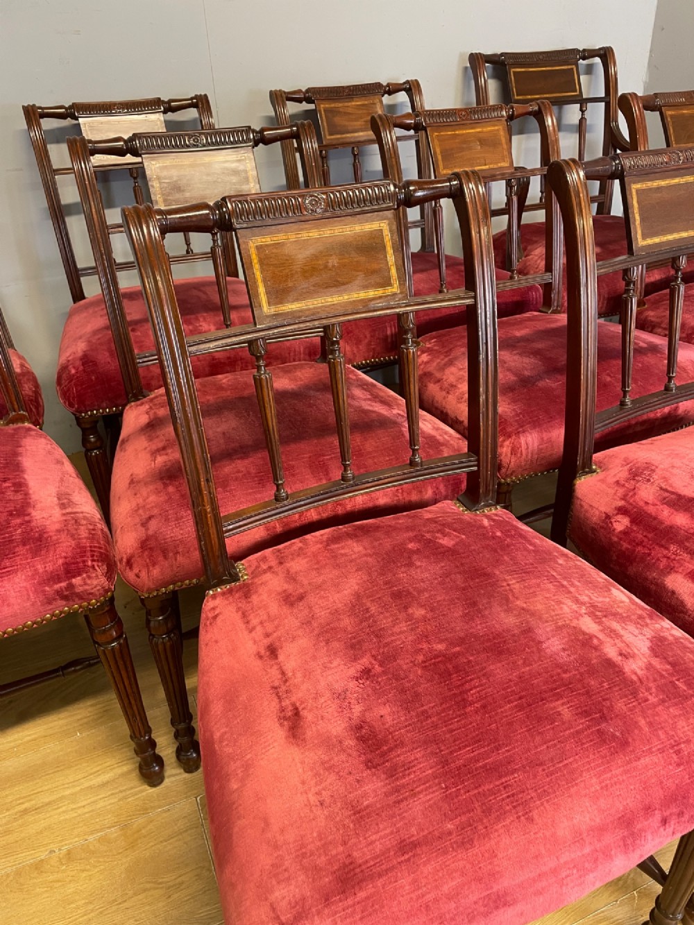 A set of ten nineteenth-century mahogany dining chairs featuring rectangular inlaid backrests, spindle uprights, and red velvet upholstered seats with brass stud detailing. View IV