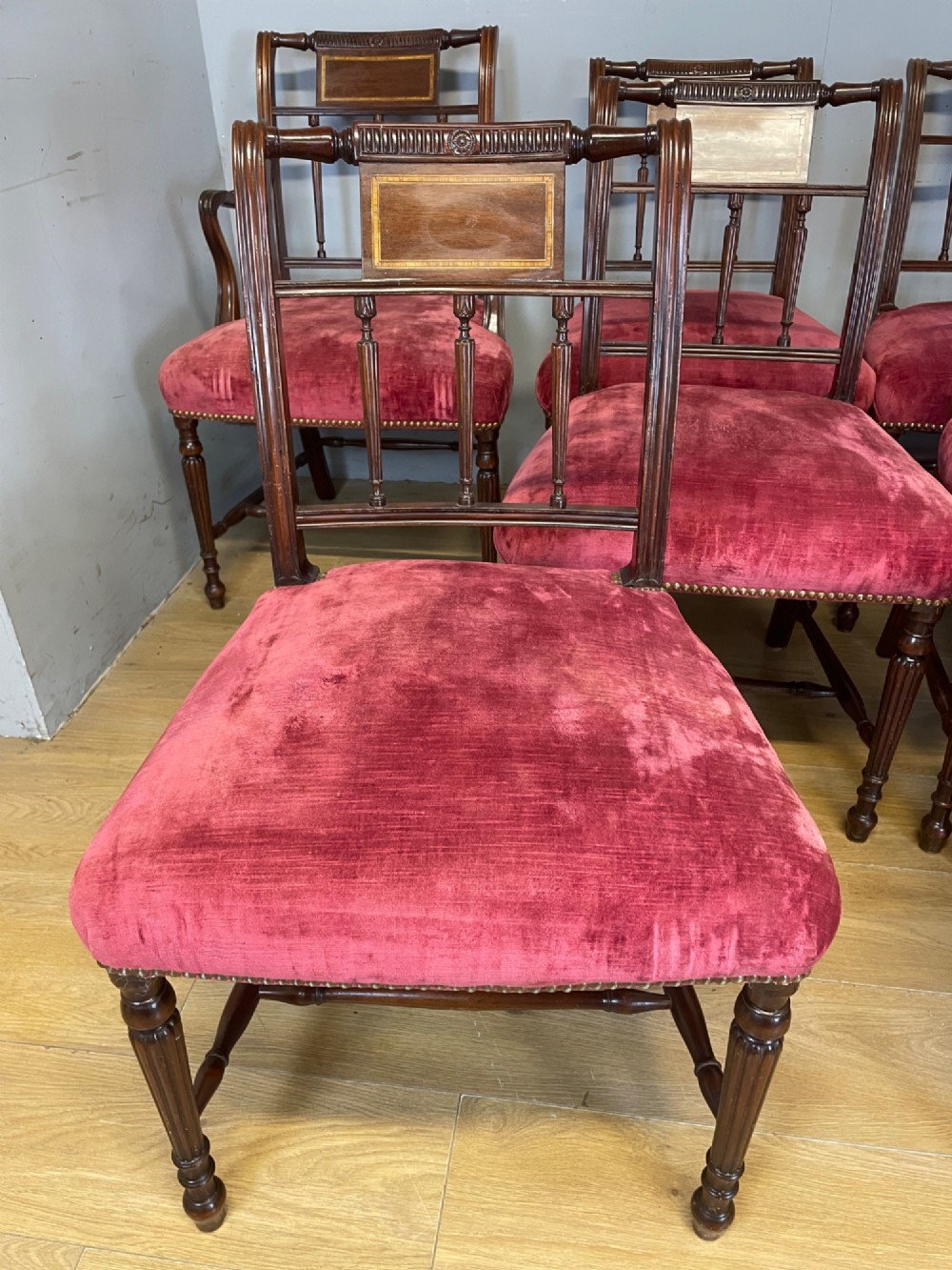 A set of ten nineteenth-century mahogany dining chairs featuring rectangular inlaid backrests, spindle uprights, and red velvet upholstered seats with brass stud detailing. View IX