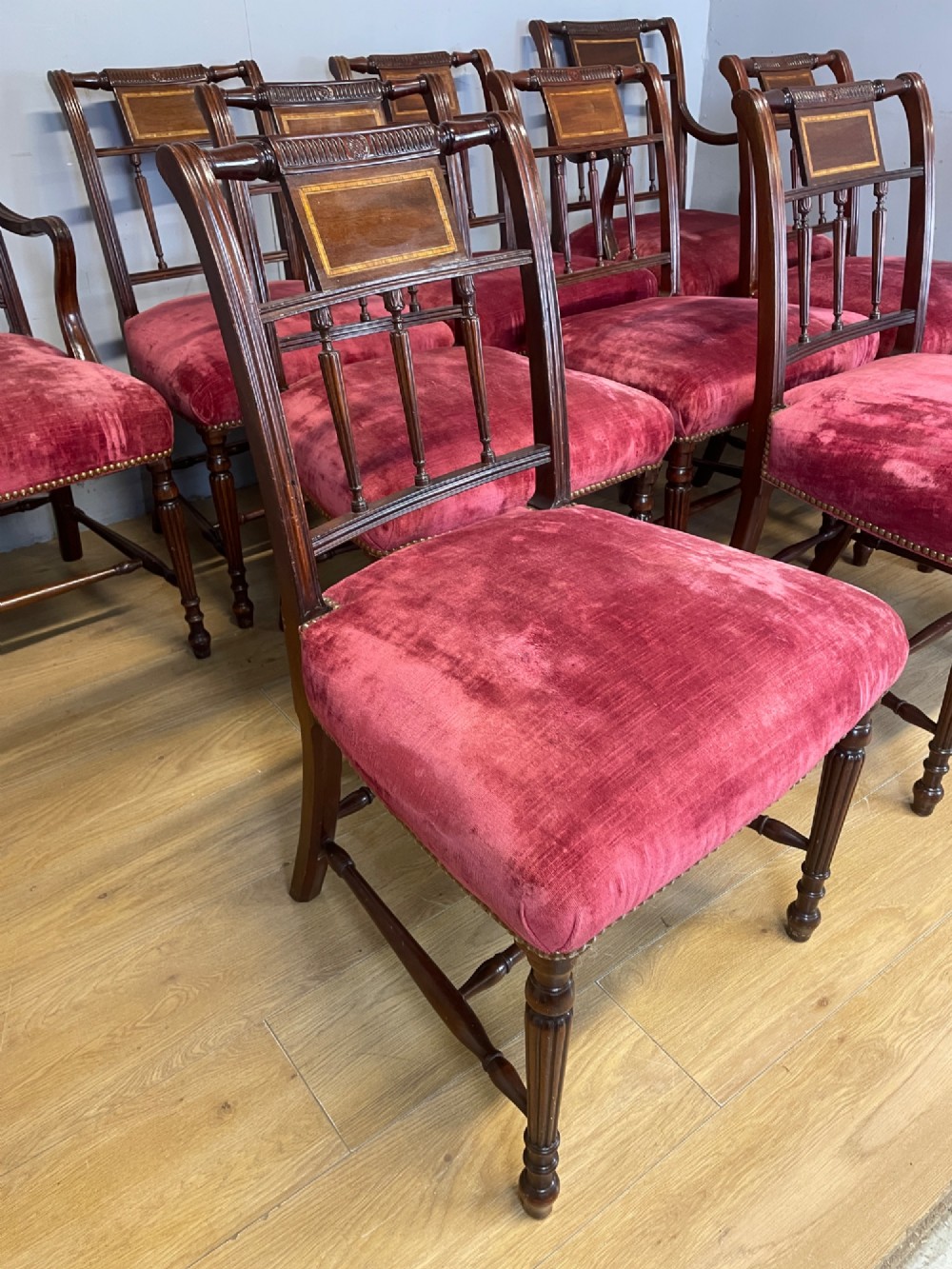 A set of ten nineteenth-century mahogany dining chairs featuring rectangular inlaid backrests, spindle uprights, and red velvet upholstered seats with brass stud detailing. View X