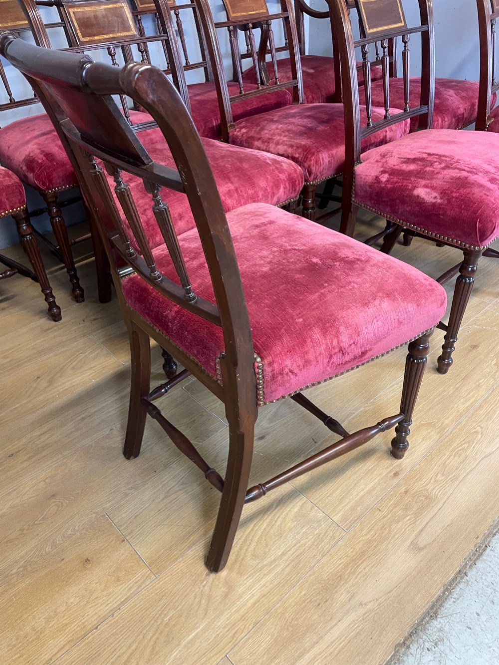 A set of ten nineteenth-century mahogany dining chairs featuring rectangular inlaid backrests, spindle uprights, and red velvet upholstered seats with brass stud detailing. View XII