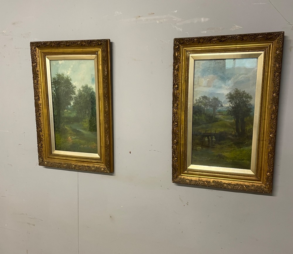 A pair of vertical late 19th-century oil paintings depicting green river and woodland landscapes in ornate gilded frames.
