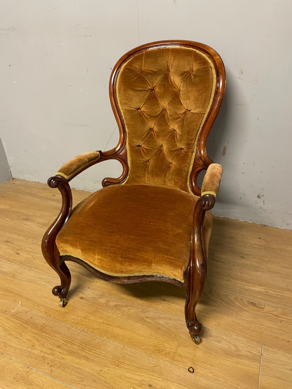 A Victorian walnut spoon back armchair featuring button-tufted mustard velvet upholstery, open carved armrests, and cabriole legs on brass castors.