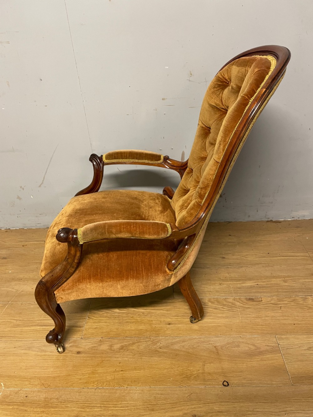 A Victorian walnut spoon back armchair featuring button-tufted mustard velvet upholstery, open carved armrests, and cabriole legs on brass castors. View VI