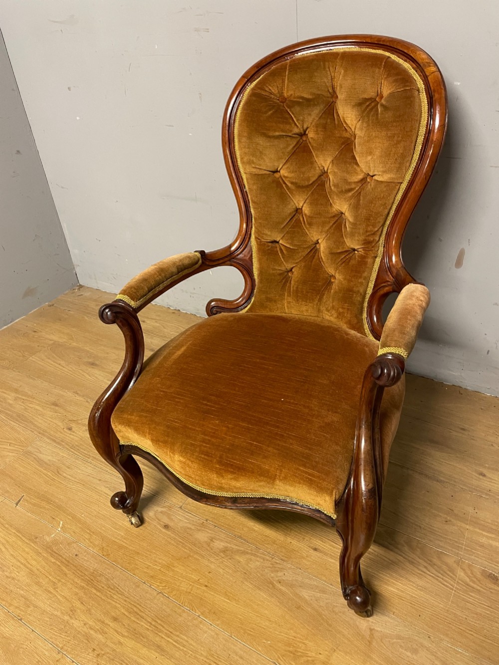 A Victorian walnut spoon back armchair featuring button-tufted mustard velvet upholstery, open carved armrests, and cabriole legs on brass castors. View X