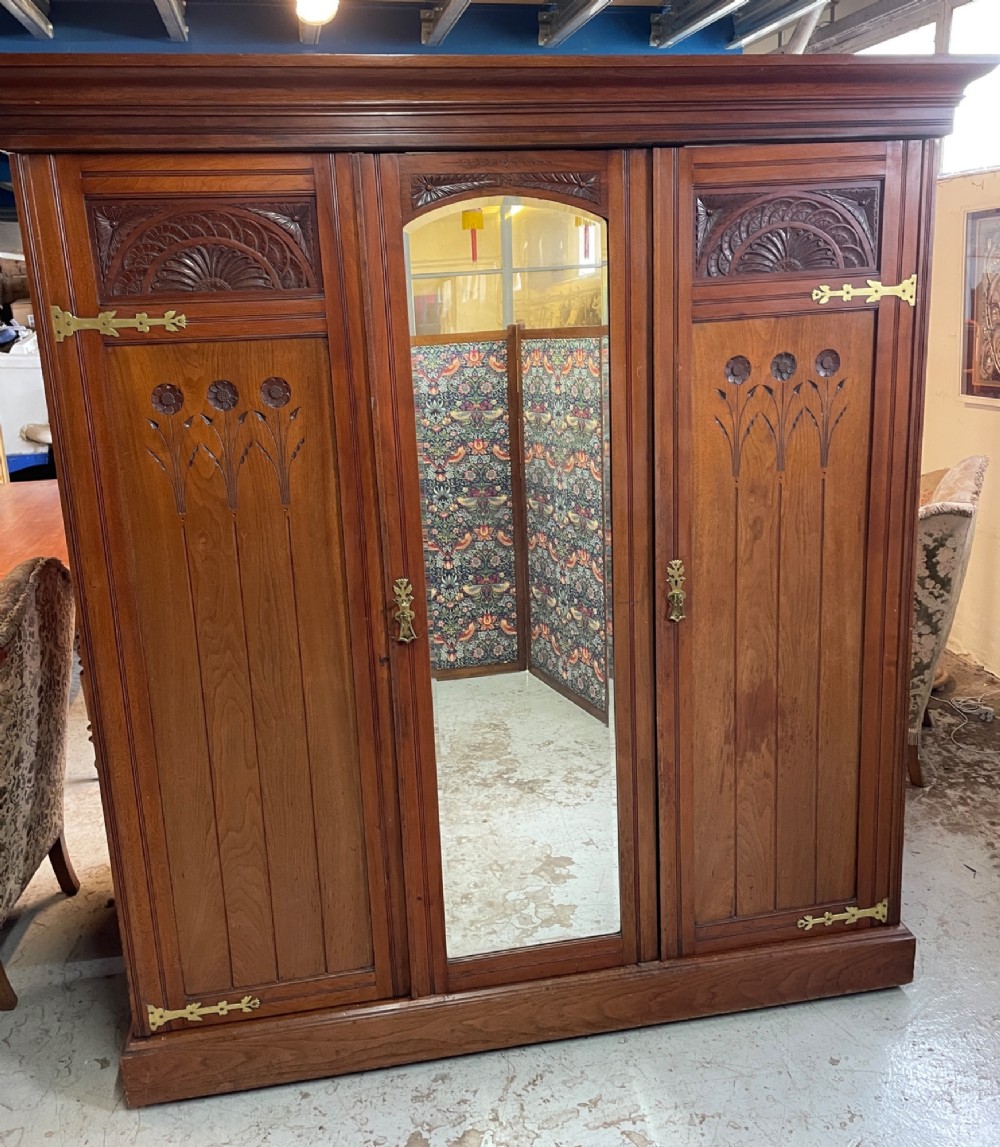 An Arts and Crafts walnut triple wardrobe featuring two outer timber doors with carved stylised flower panels and a central arched mirrored door with decorative brass hinges.
