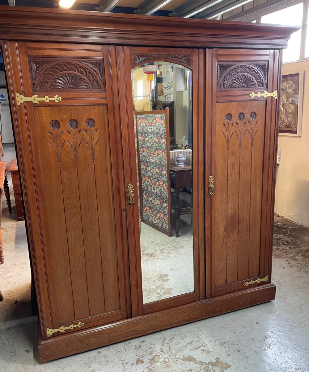 An Arts and Crafts walnut triple wardrobe featuring two outer timber doors with carved stylised flower panels and a central arched mirrored door with decorative brass hinges. View II