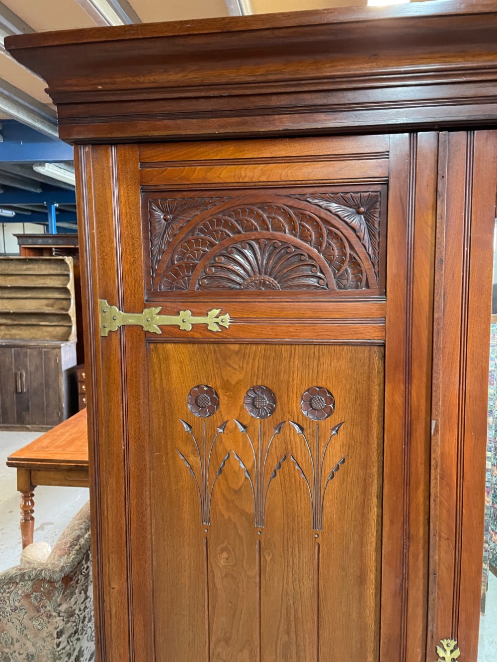 An Arts and Crafts walnut triple wardrobe featuring two outer timber doors with carved stylised flower panels and a central arched mirrored door with decorative brass hinges. View III