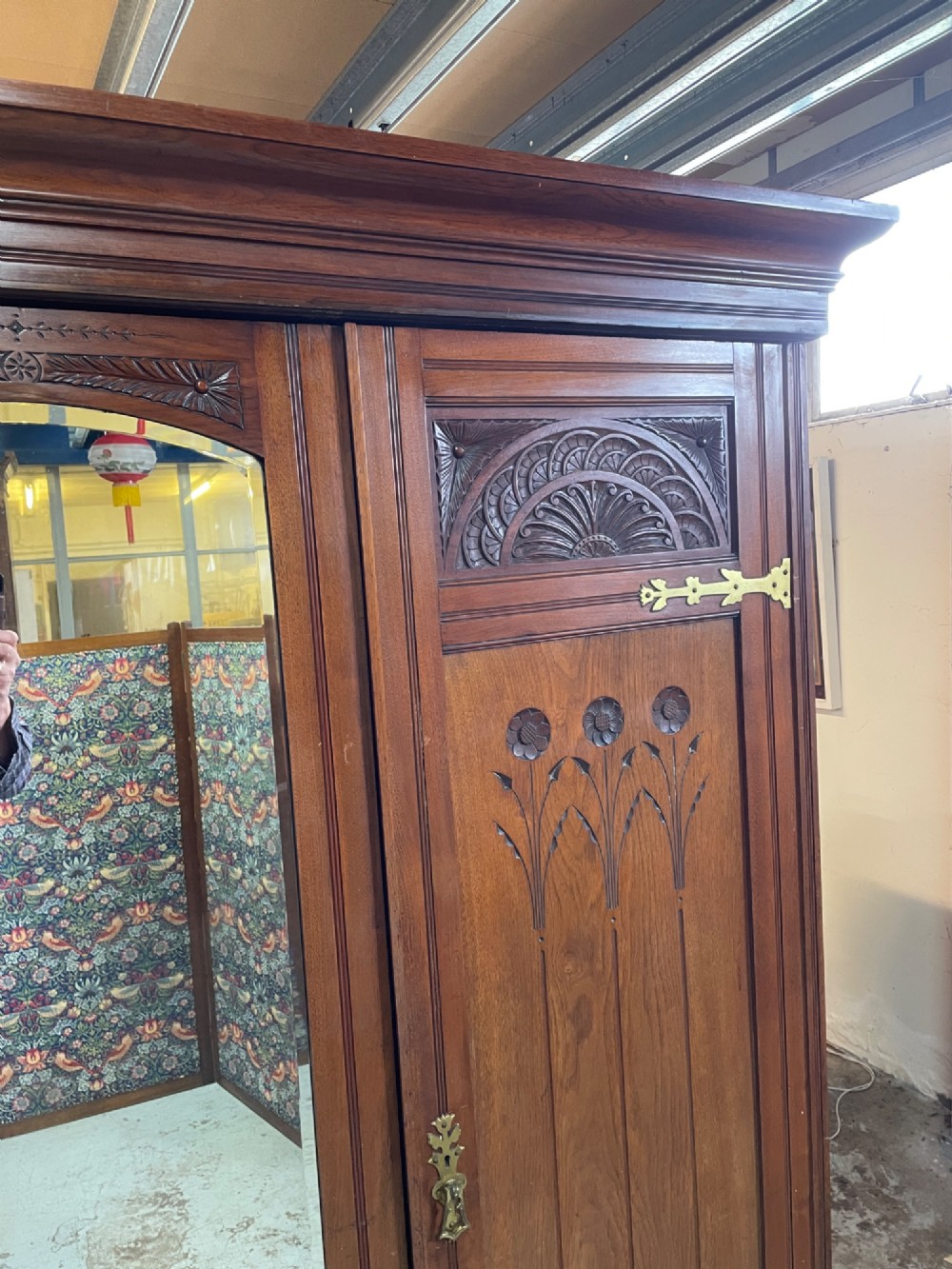 An Arts and Crafts walnut triple wardrobe featuring two outer timber doors with carved stylised flower panels and a central arched mirrored door with decorative brass hinges. View IV