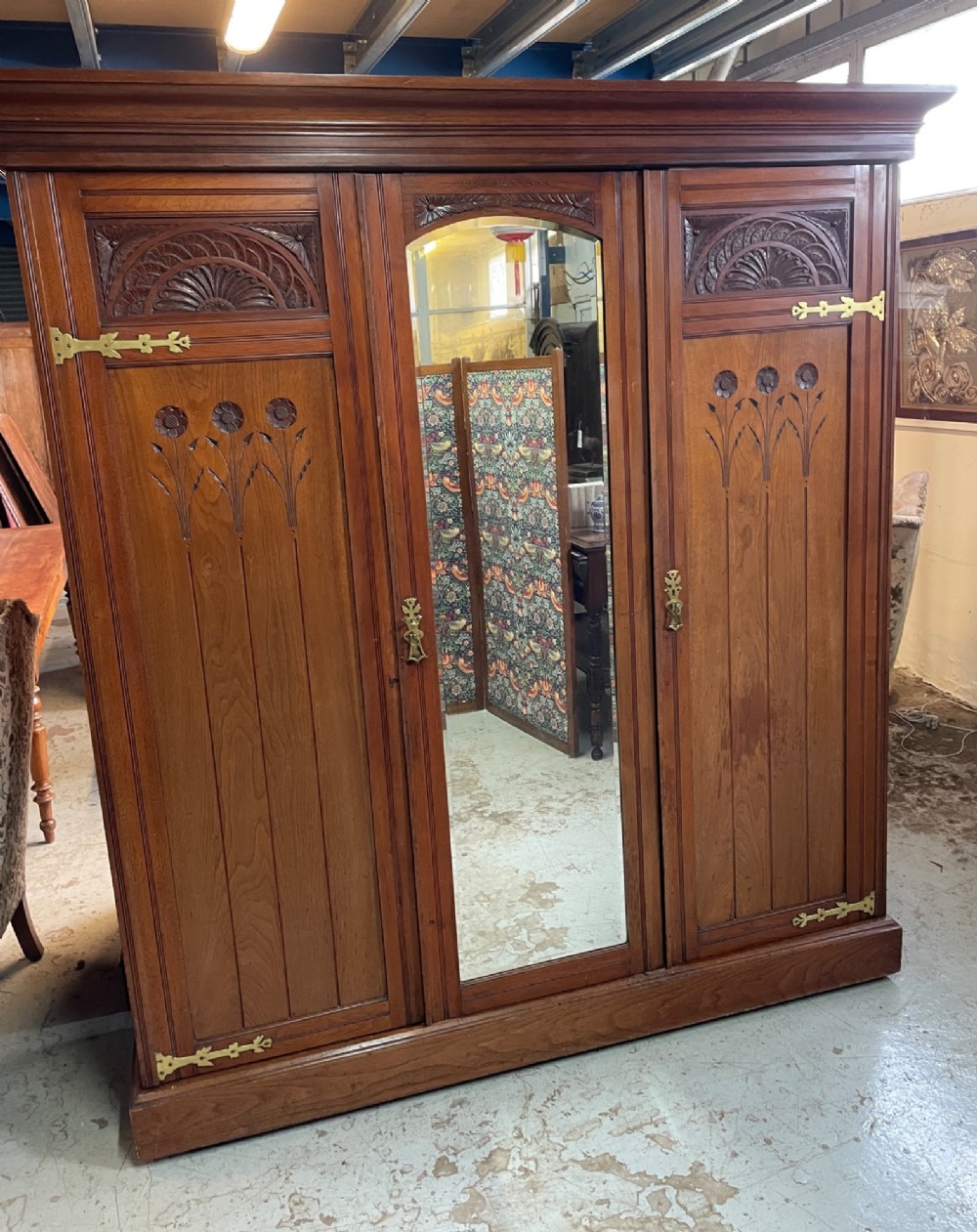 An Arts and Crafts walnut triple wardrobe featuring two outer timber doors with carved stylised flower panels and a central arched mirrored door with decorative brass hinges. View V