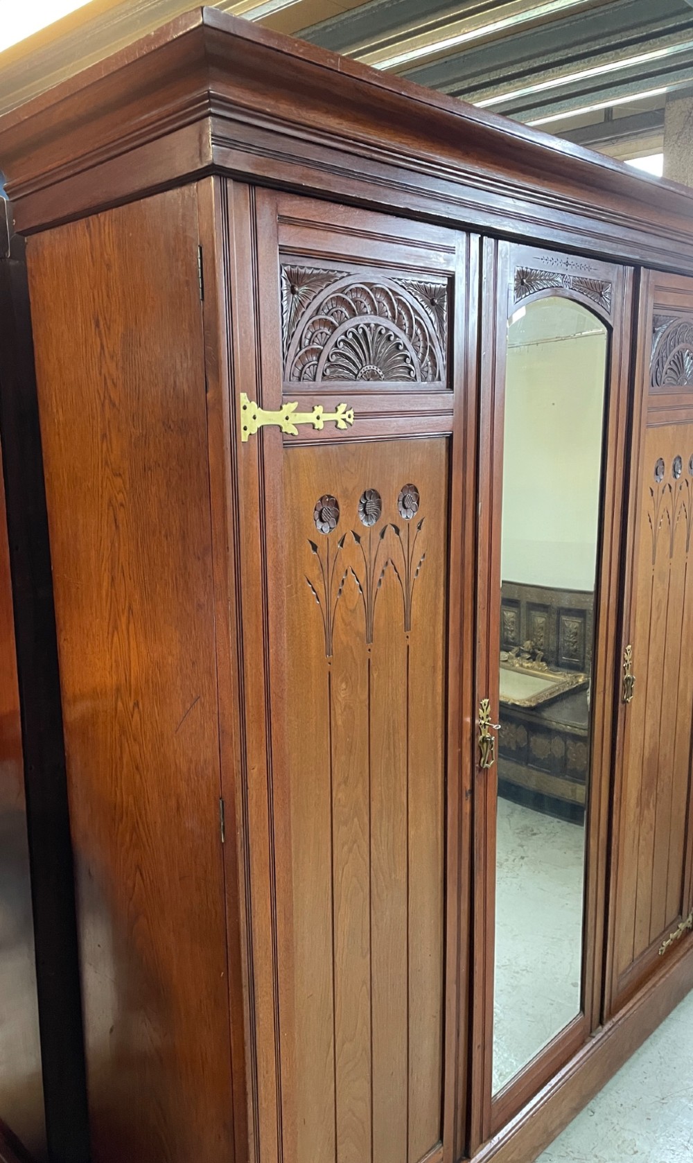 An Arts and Crafts walnut triple wardrobe featuring two outer timber doors with carved stylised flower panels and a central arched mirrored door with decorative brass hinges. View VI