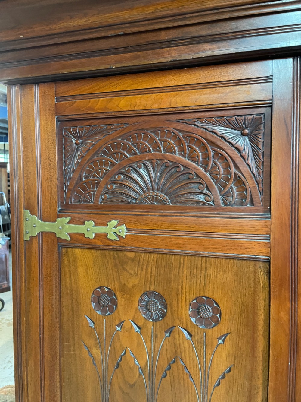 An Arts and Crafts walnut triple wardrobe featuring two outer timber doors with carved stylised flower panels and a central arched mirrored door with decorative brass hinges. View VII