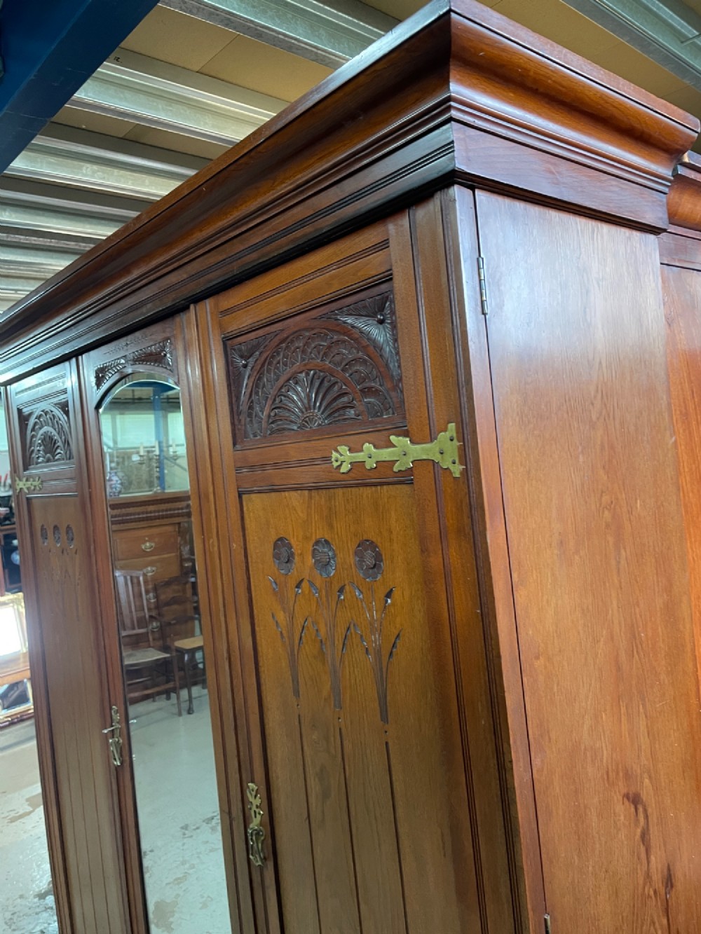 An Arts and Crafts walnut triple wardrobe featuring two outer timber doors with carved stylised flower panels and a central arched mirrored door with decorative brass hinges. View VIII