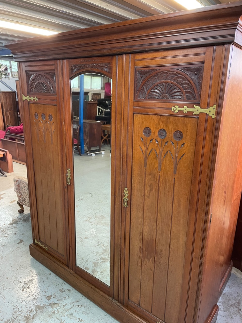 An Arts and Crafts walnut triple wardrobe featuring two outer timber doors with carved stylised flower panels and a central arched mirrored door with decorative brass hinges. View XIV