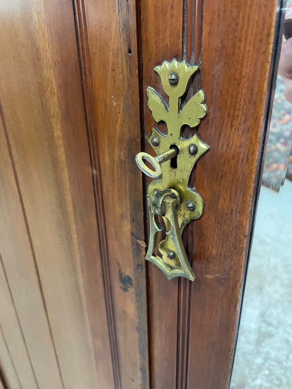 An Arts and Crafts walnut triple wardrobe featuring two outer timber doors with carved stylised flower panels and a central arched mirrored door with decorative brass hinges. View XV