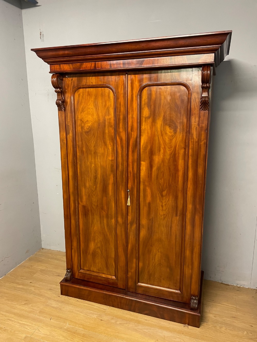 A 19th-century double wardrobe constructed from flame mahogany featuring two arched panelled doors, carved corbel accents, a moulded cornice, and a flat plinth base.