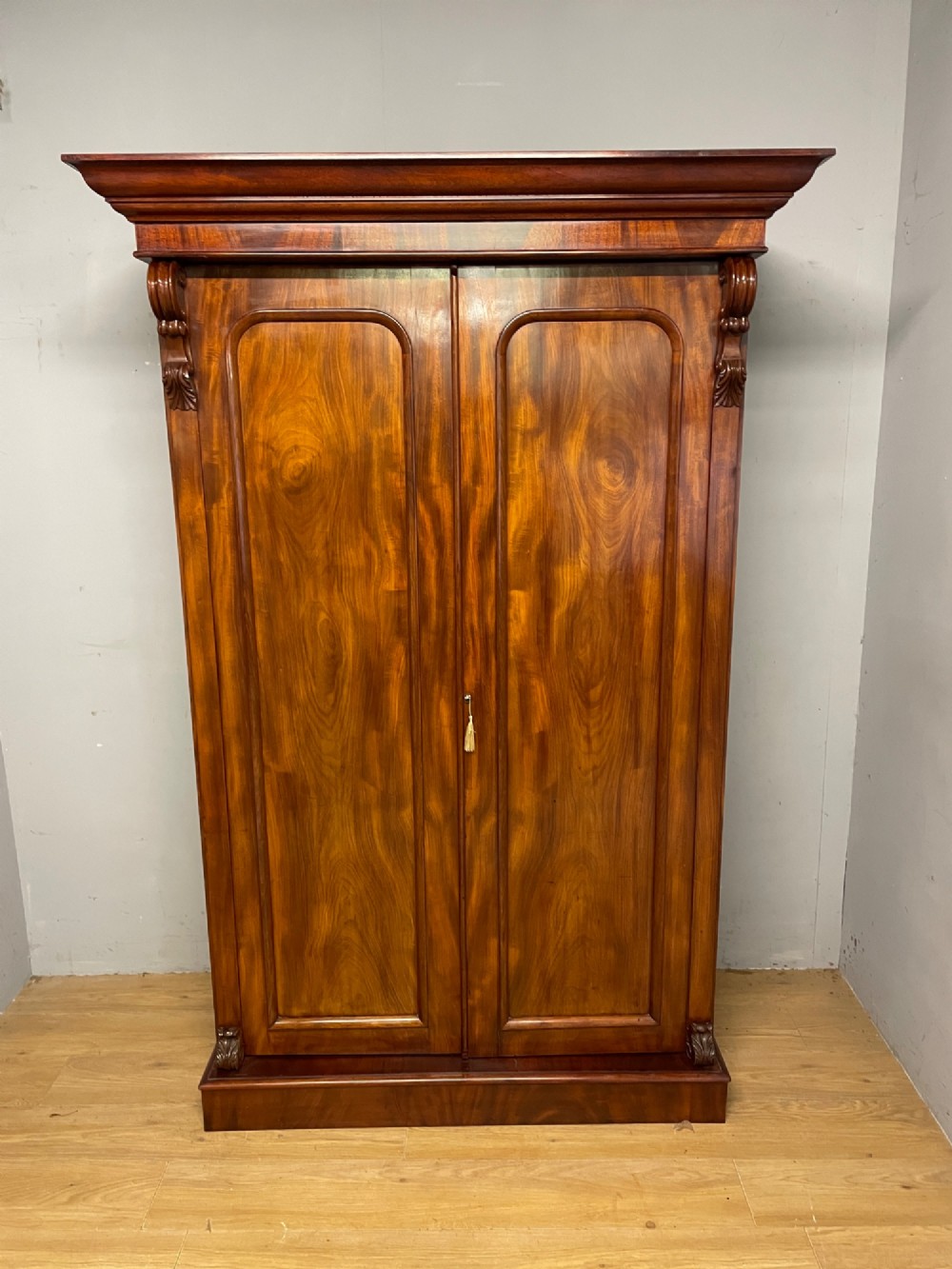 A 19th-century double wardrobe constructed from flame mahogany featuring two arched panelled doors, carved corbel accents, a moulded cornice, and a flat plinth base. View III