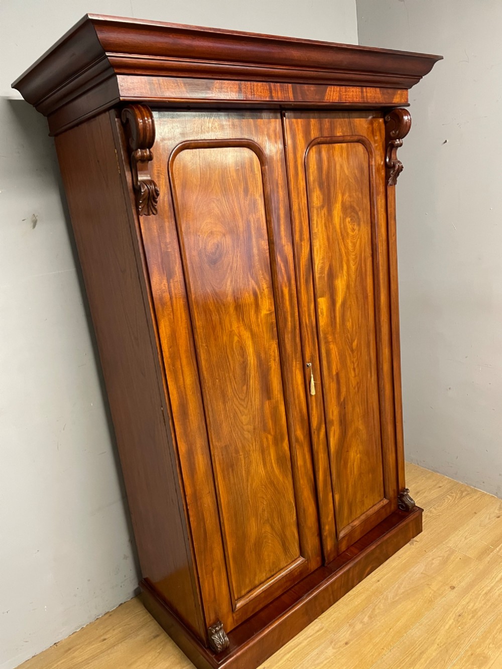 A 19th-century double wardrobe constructed from flame mahogany featuring two arched panelled doors, carved corbel accents, a moulded cornice, and a flat plinth base. View IV