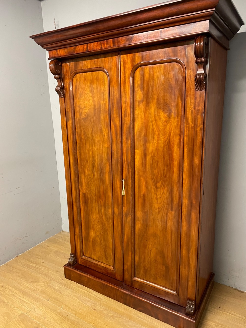 A 19th-century double wardrobe constructed from flame mahogany featuring two arched panelled doors, carved corbel accents, a moulded cornice, and a flat plinth base. View XIII