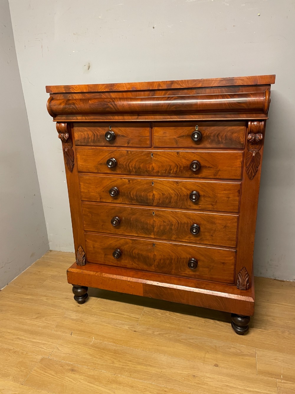 A 19th-century Scottish flame mahogany chest of drawers featuring a top cushion drawer, carved corbels, five lower drawers with turned wooden knobs, and turned feet.