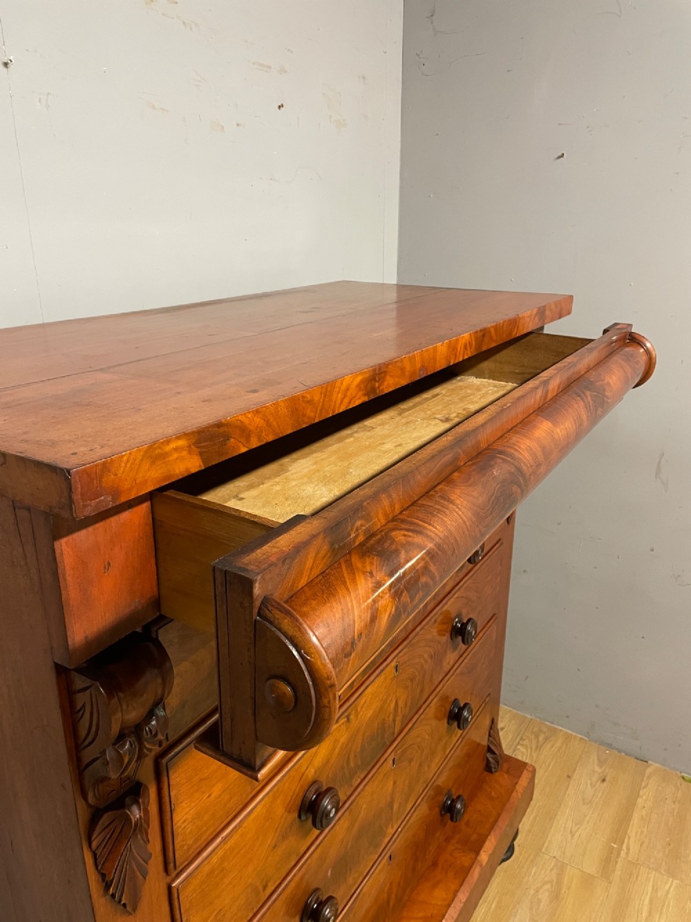 A 19th-century Scottish flame mahogany chest of drawers featuring a top cushion drawer, carved corbels, five lower drawers with turned wooden knobs, and turned feet. View V