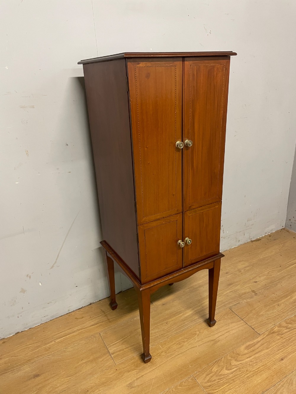 A tall slim Edwardian mahogany cabinet featuring four doors with line inlay, brass knobs, and tall tapered legs.