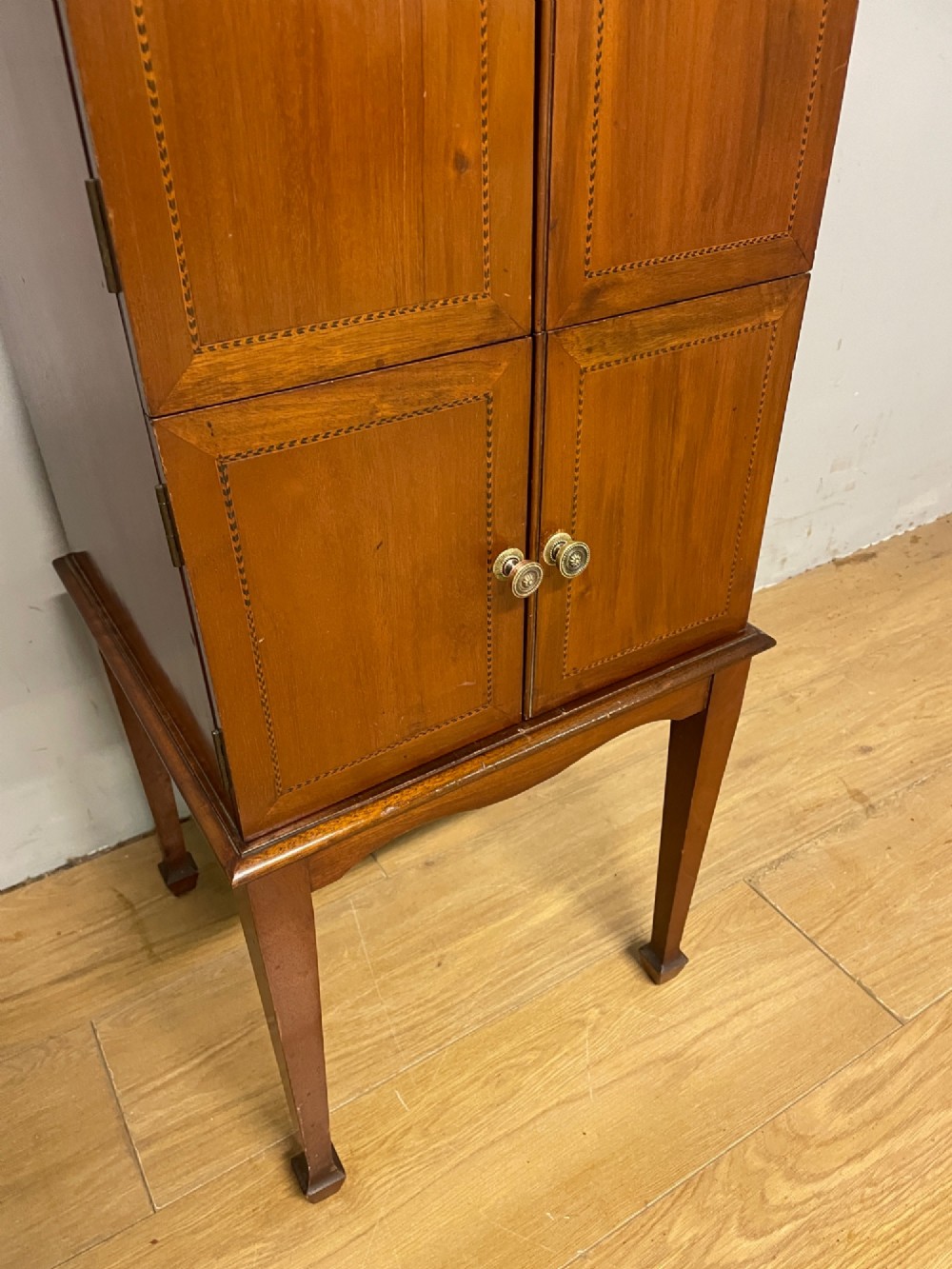 A tall slim Edwardian mahogany cabinet featuring four doors with line inlay, brass knobs, and tall tapered legs. View V