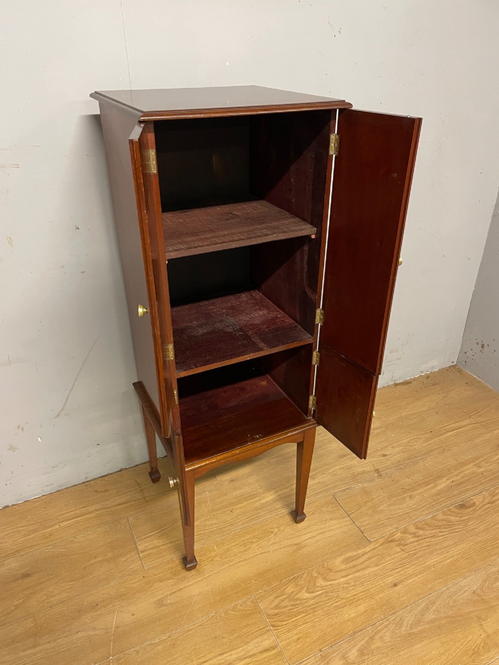 A tall slim Edwardian mahogany cabinet featuring four doors with line inlay, brass knobs, and tall tapered legs. View VI