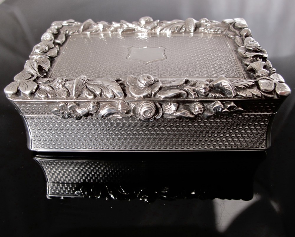 A rectangular sterling silver tabletop box featuring intricate engine-turned geometric patterns, a raised floral border, and an unengraved shield cartouche in the centre.