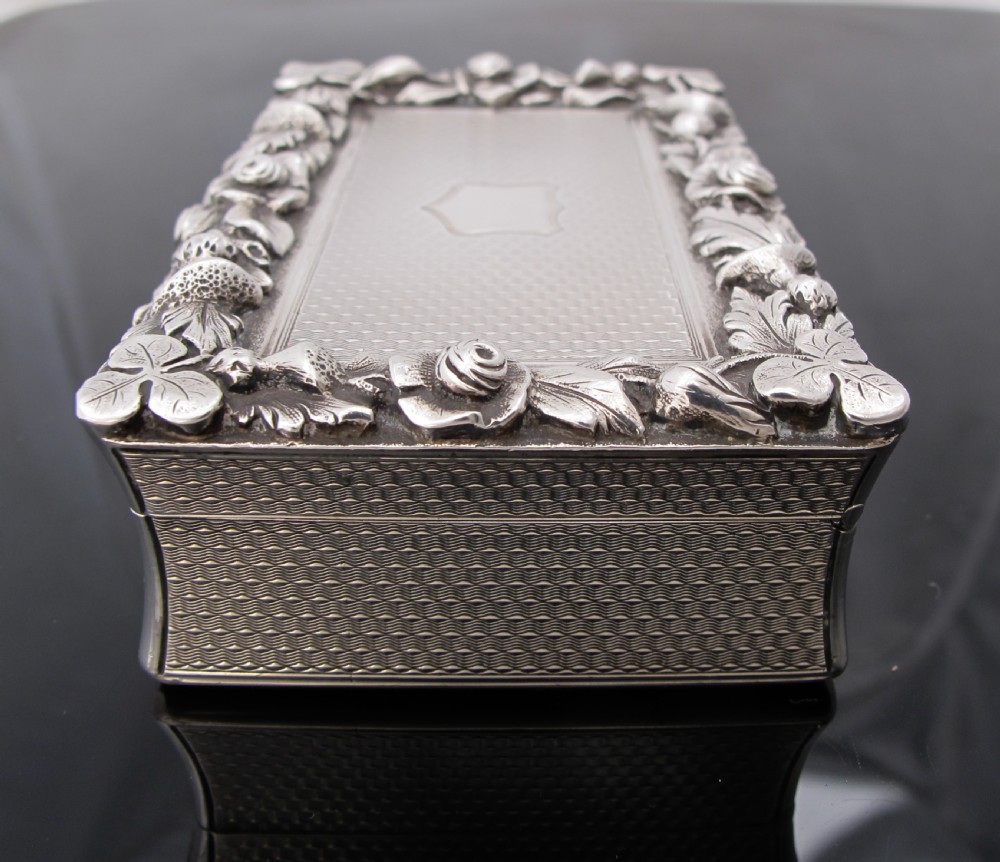 A rectangular sterling silver tabletop box featuring intricate engine-turned geometric patterns, a raised floral border, and an unengraved shield cartouche in the centre. View II