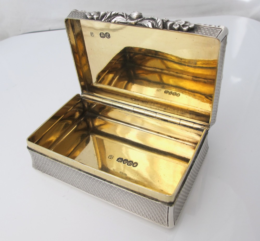 A rectangular sterling silver tabletop box featuring intricate engine-turned geometric patterns, a raised floral border, and an unengraved shield cartouche in the centre. View III