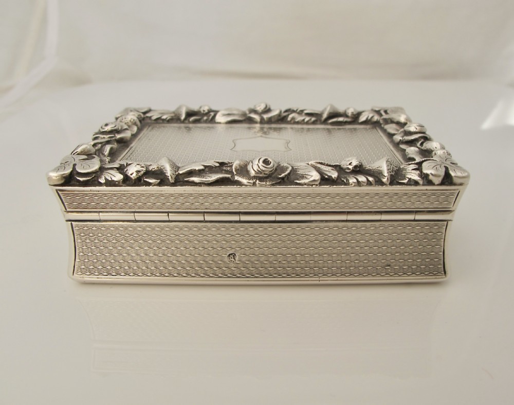 A rectangular sterling silver tabletop box featuring intricate engine-turned geometric patterns, a raised floral border, and an unengraved shield cartouche in the centre. View VI