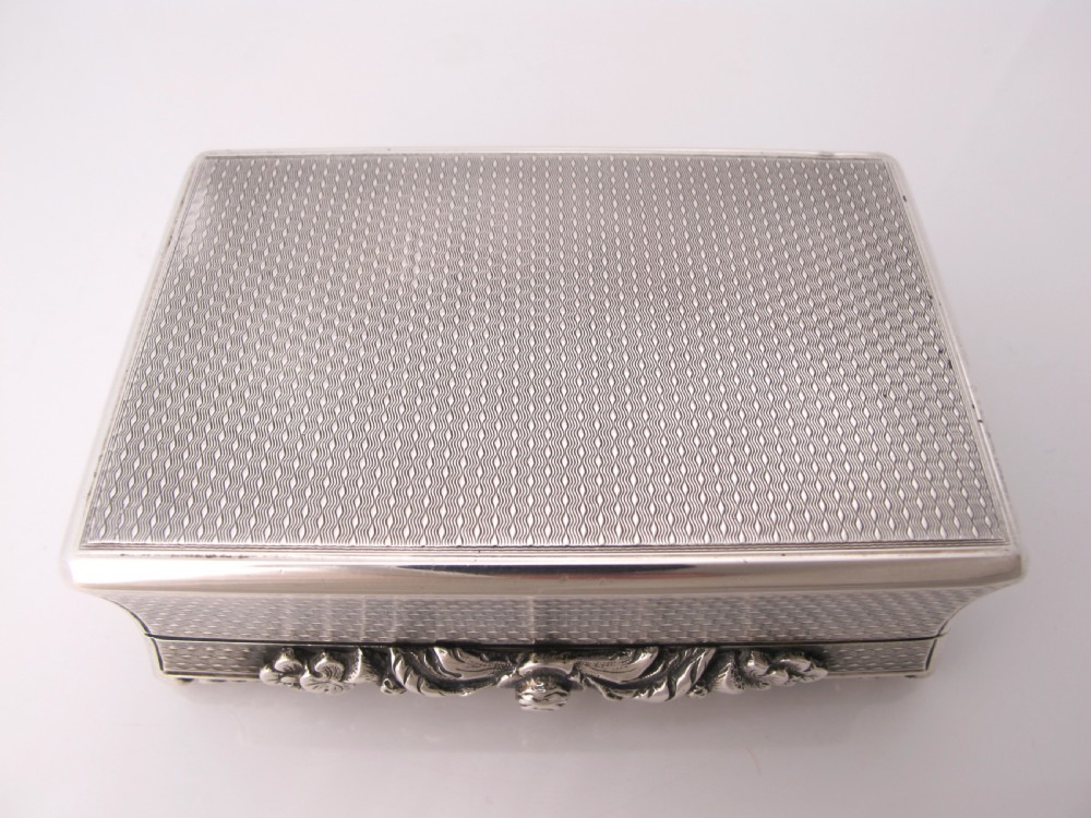 A rectangular sterling silver tabletop box featuring intricate engine-turned geometric patterns, a raised floral border, and an unengraved shield cartouche in the centre. View VII
