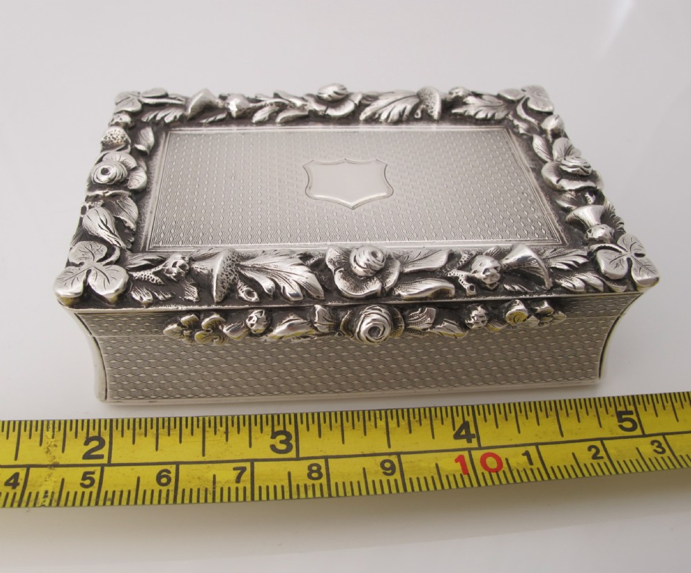 A rectangular sterling silver tabletop box featuring intricate engine-turned geometric patterns, a raised floral border, and an unengraved shield cartouche in the centre. View IX