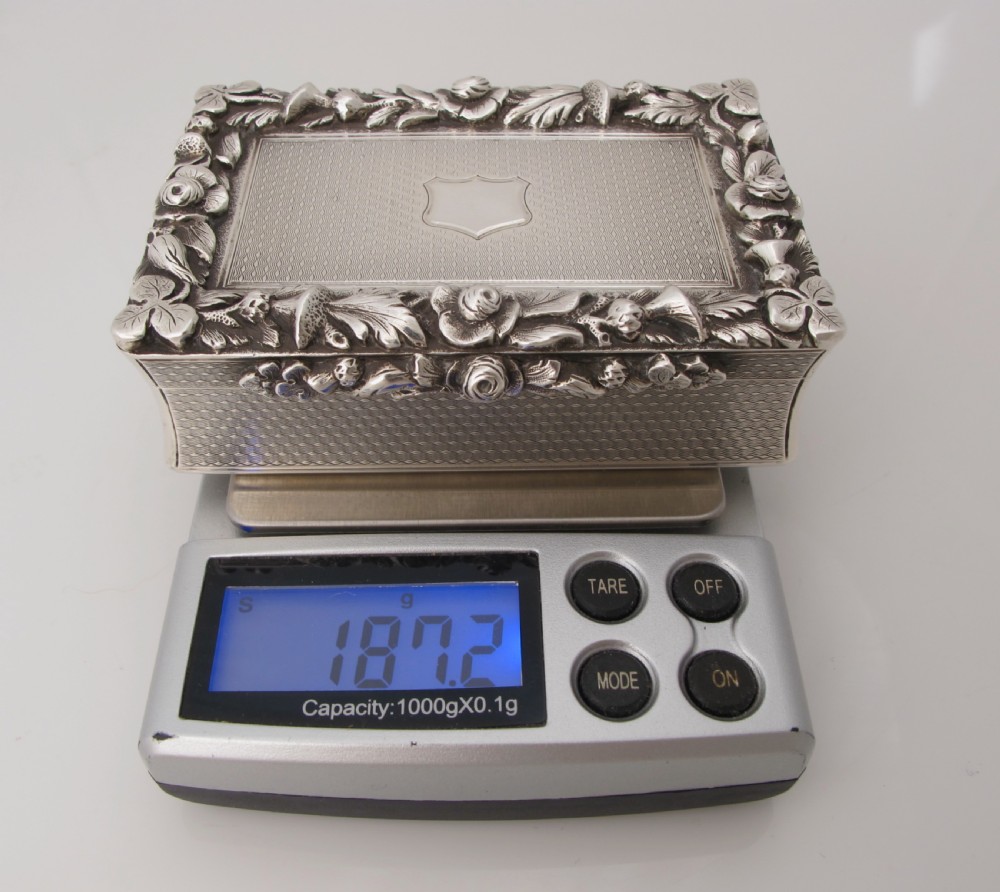 A rectangular sterling silver tabletop box featuring intricate engine-turned geometric patterns, a raised floral border, and an unengraved shield cartouche in the centre. View X