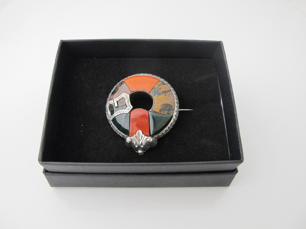 A circular Victorian Scottish silver brooch inlaid with segmented orange, green, and brown agate stones featuring an engraved silver buckle motif.