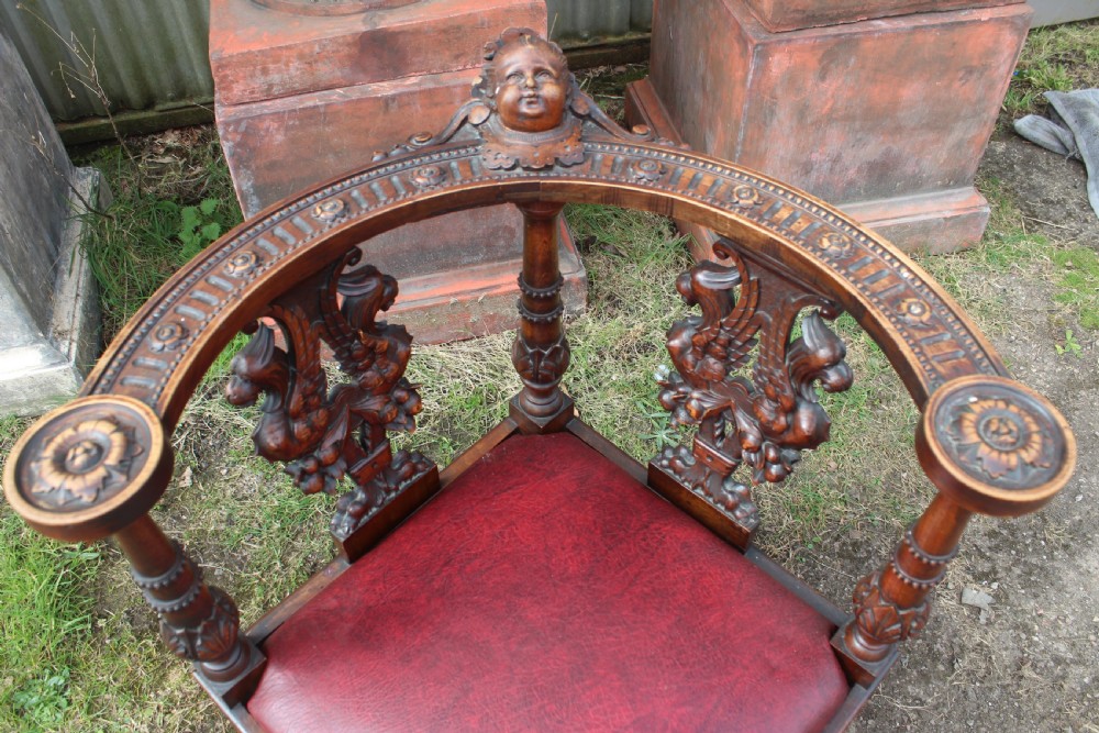 A carved dark wood corner chair featuring a red leather seat, carved splats depicting winged creatures, turned legs, and a cherub motif on the top rail. View III