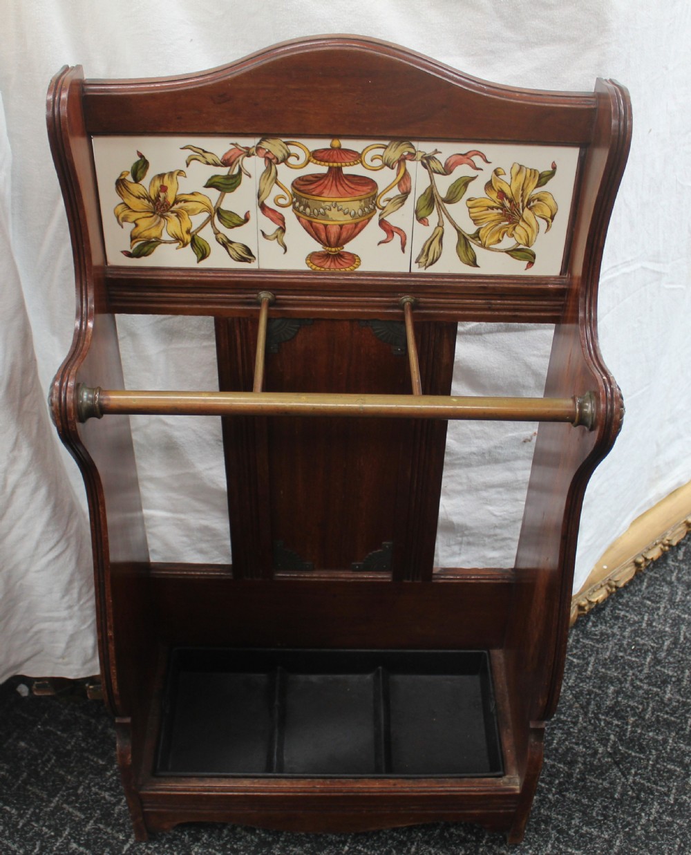 A late Victorian dark oak stick stand featuring a brass retaining bar, ceramic tile insert decorated with yellow lilies and an urn motif, and a removable black cast iron drip tray at the base.