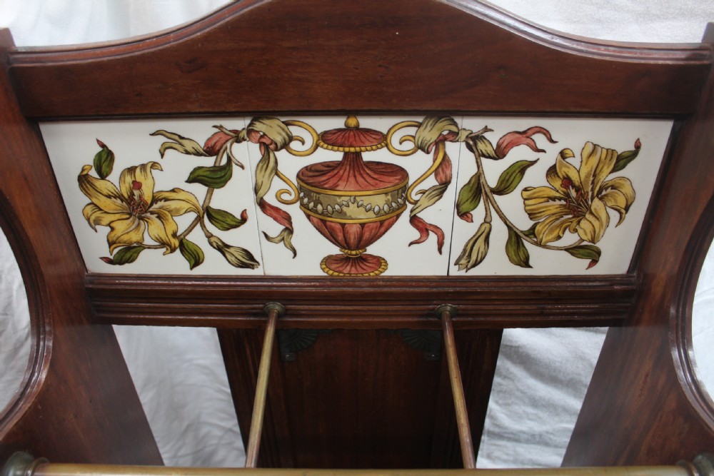A late Victorian dark oak stick stand featuring a brass retaining bar, ceramic tile insert decorated with yellow lilies and an urn motif, and a removable black cast iron drip tray at the base. View III
