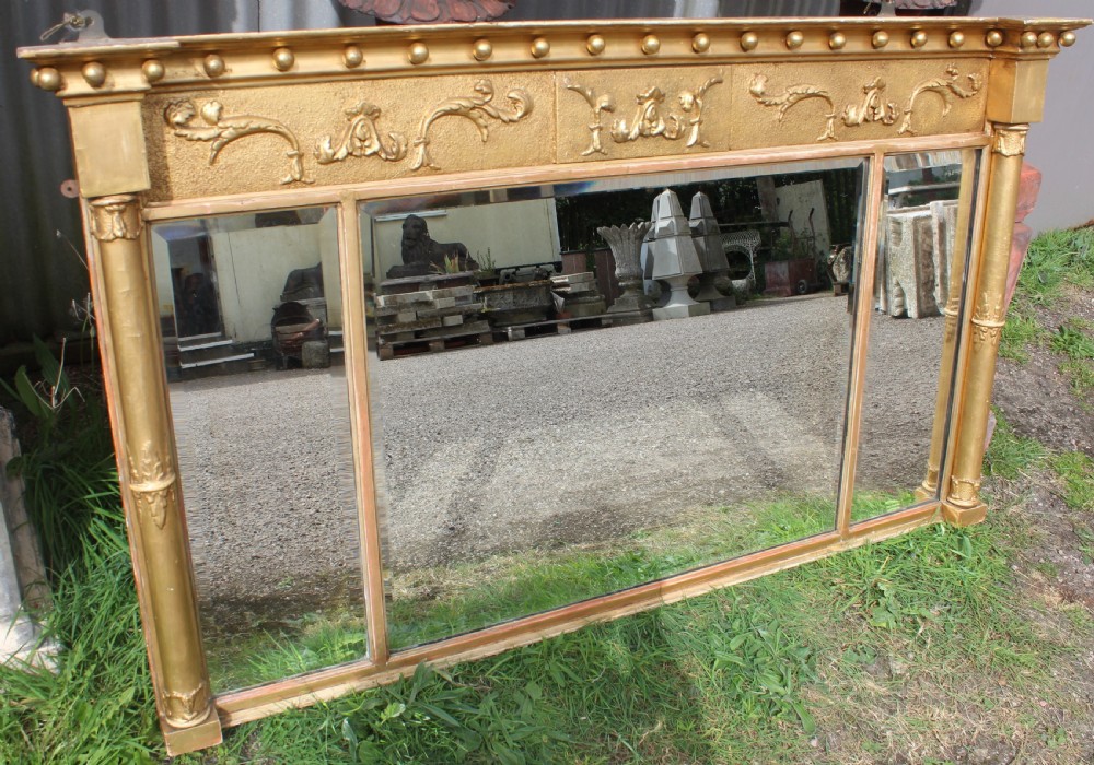 A wide gilded landscape overmantel mirror featuring three glass panels flanked by fluted columns and topped with a decorative foliate frieze.