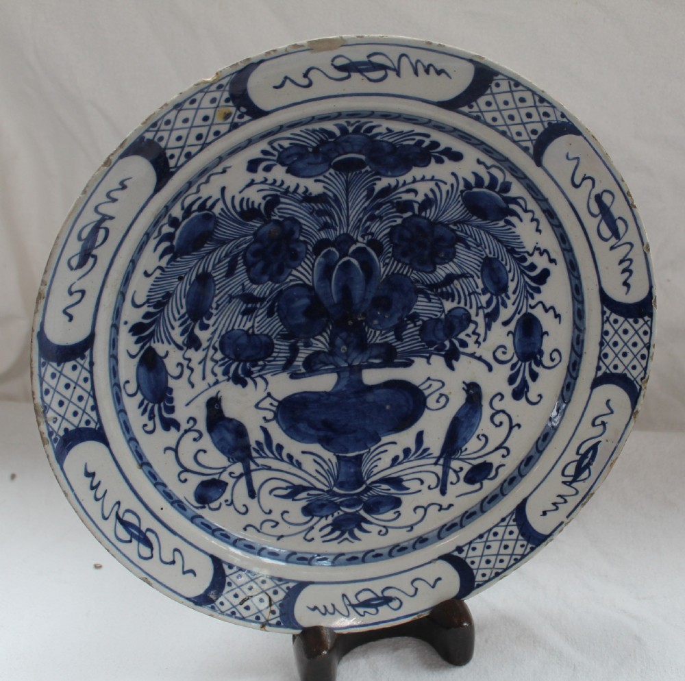 A circular tin-glazed earthenware plate featuring hand-painted cobalt blue decoration depicting a floral vase flanked by two birds within a patterned rim.