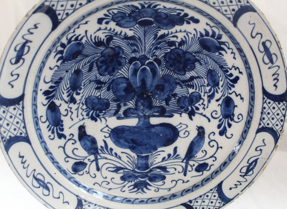 A circular tin-glazed earthenware plate featuring hand-painted cobalt blue decoration depicting a floral vase flanked by two birds within a patterned rim. View II