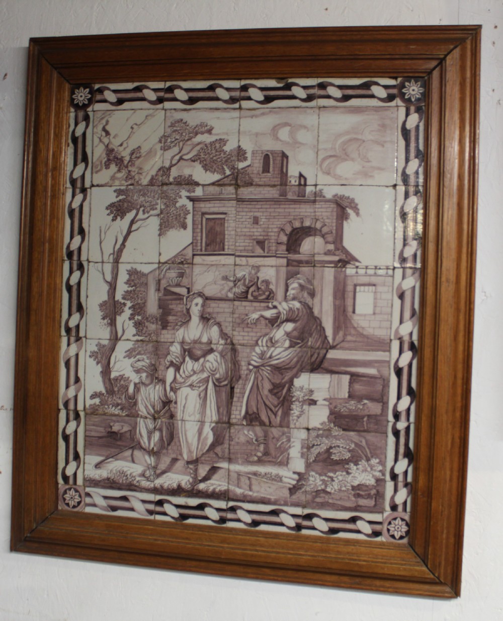 A framed multi-tile ceramic panel hand-painted in manganese monochrome tones featuring a classical scene with figures in front of architectural ruins.