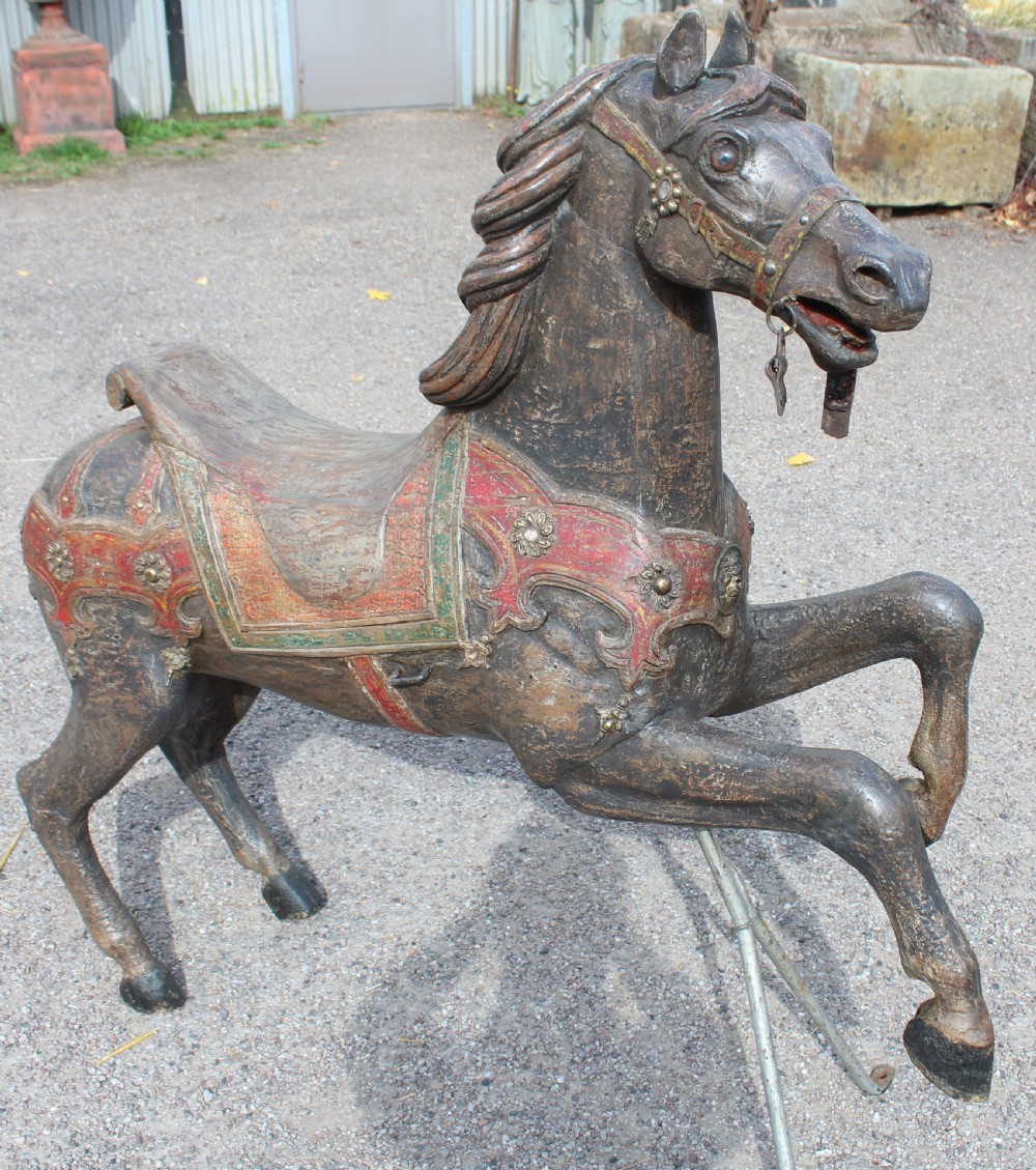 A hand-carved wooden carousel horse featuring weathered dark paint, decorative red and green saddle details, and a raised front leg pose.