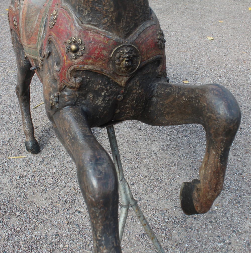 A hand-carved wooden carousel horse featuring weathered dark paint, decorative red and green saddle details, and a raised front leg pose. View IV