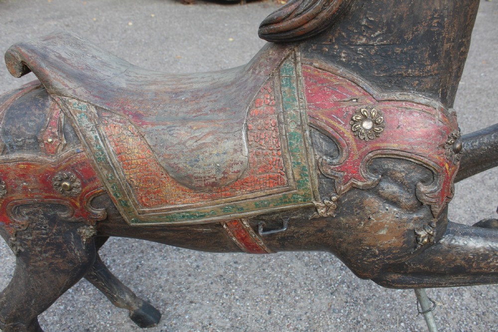 A hand-carved wooden carousel horse featuring weathered dark paint, decorative red and green saddle details, and a raised front leg pose. View V