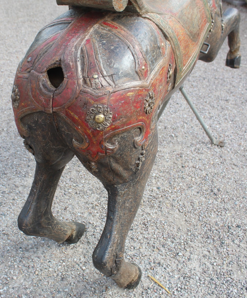 A hand-carved wooden carousel horse featuring weathered dark paint, decorative red and green saddle details, and a raised front leg pose. View VI