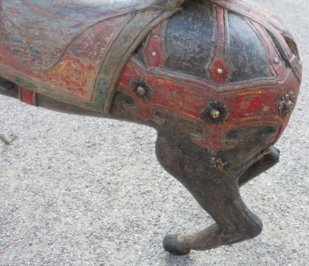 A hand-carved wooden carousel horse featuring weathered dark paint, decorative red and green saddle details, and a raised front leg pose. View VII