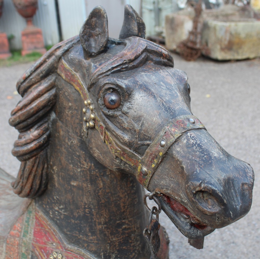 A hand-carved wooden carousel horse featuring weathered dark paint, decorative red and green saddle details, and a raised front leg pose. View X